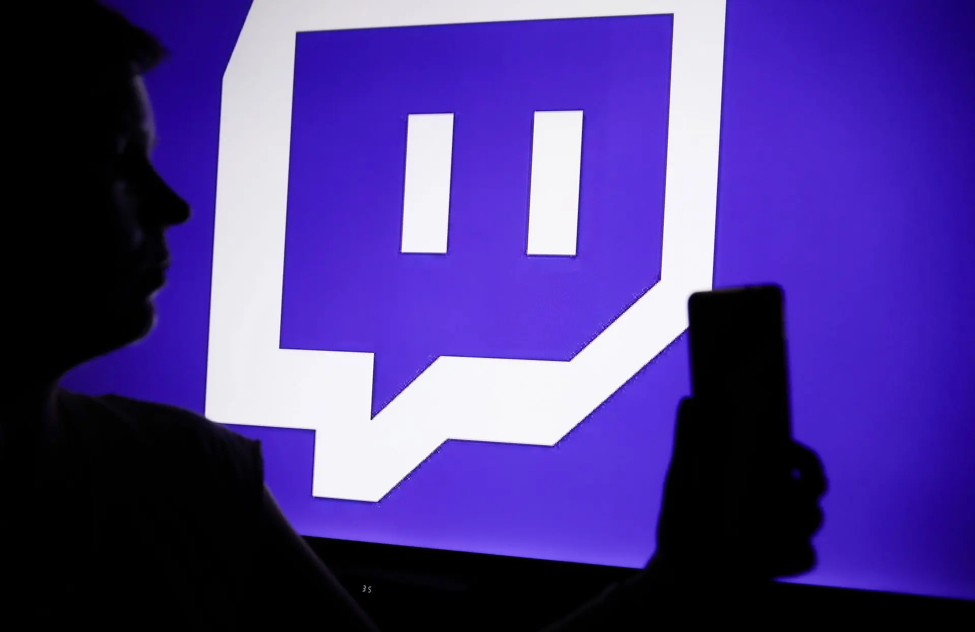 The Twitch logo with a person holding a phone in their hand (Image via Getty)