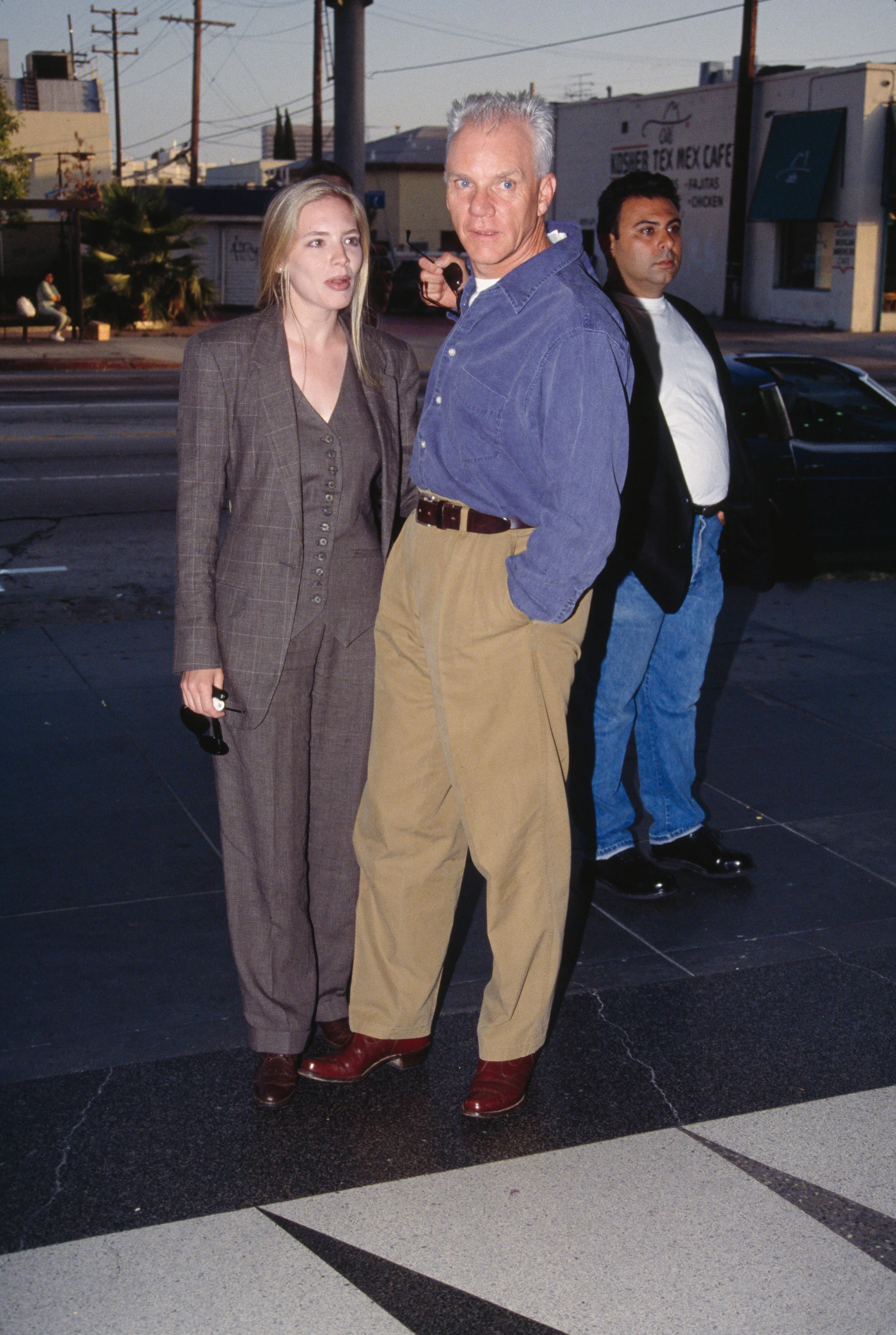 British actor Malcolm McDowell, wear tan trousers and a blue shirt over a white t-shirt, and his wife Kelley Kuhr, wearing a grey tweed trouser suit, location unknown, circa 1995.  (Photo by Vinnie Zuffante/Getty Images)