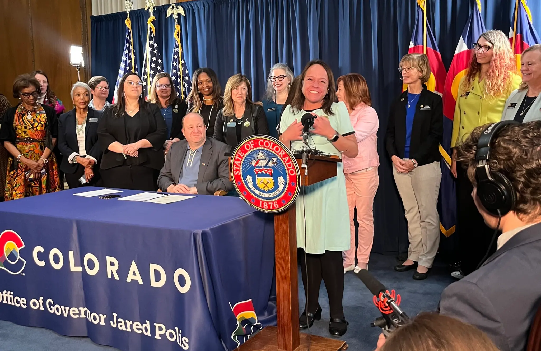 Colorado State Senator Faith Winter standing on the podium beside Governor Jared Polis (Image via Facebook/Senator Faith Winter)