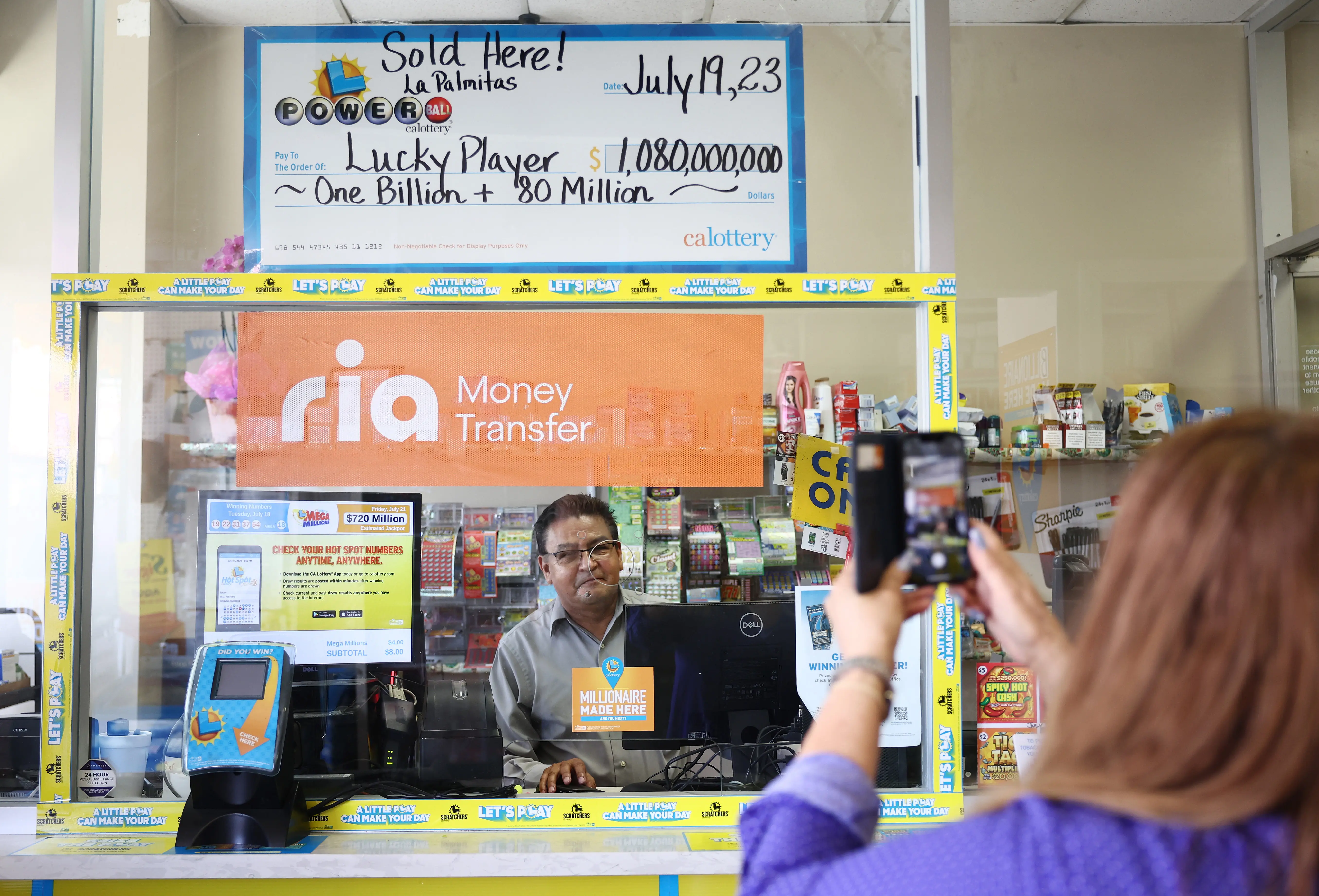 LOS ANGELES, CALIFORNIA - JULY 20: A person poses for a photo beneath an enlarged symbolic check for $1.08 billion in Las Palmitas Mini Market on July 20, 2023 in Los Angeles, California. The $1.08 billion winning Powerball ticket was sold at the Las Palmitas Mini Market for the July 19th drawing. The jackpot is the third largest in Powerball history and was picked after three months of drawings without a winner. The mini market is located in the downtown Fashion District close to Skid Row. (Photo by Mario Tama/Getty Images)