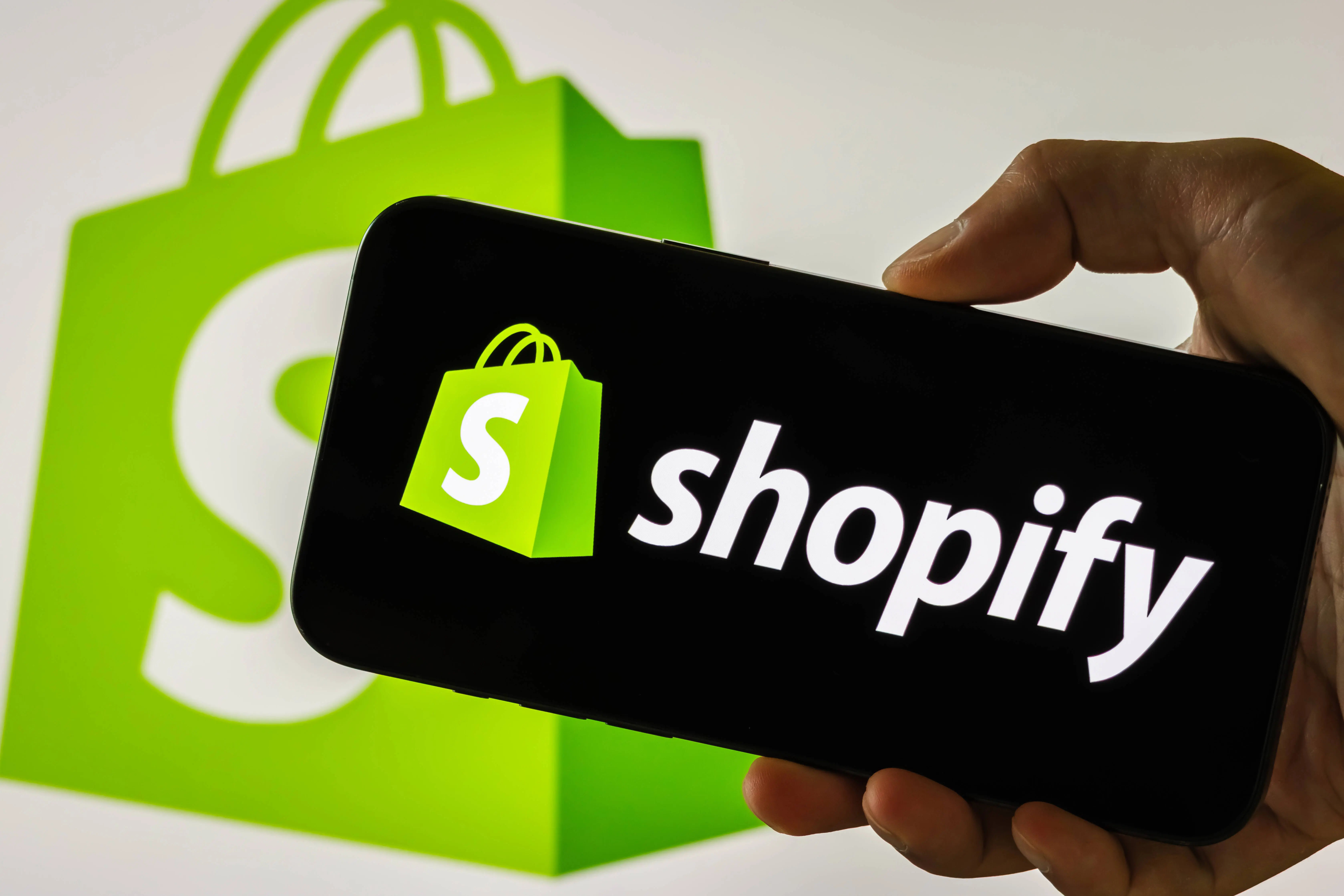 CHONGQING, CHINA - JULY 31: In this photo illustration, a person holds a smartphone displaying the logo of Shopify Inc. (NYSE: SHOP), a Canadian multinational e-commerce company, in front of a screen showing the company's green shopping bag icon, on July 31, 2025 in Chongqing, China. (Photo illustration by Cheng Xin/Getty Images)