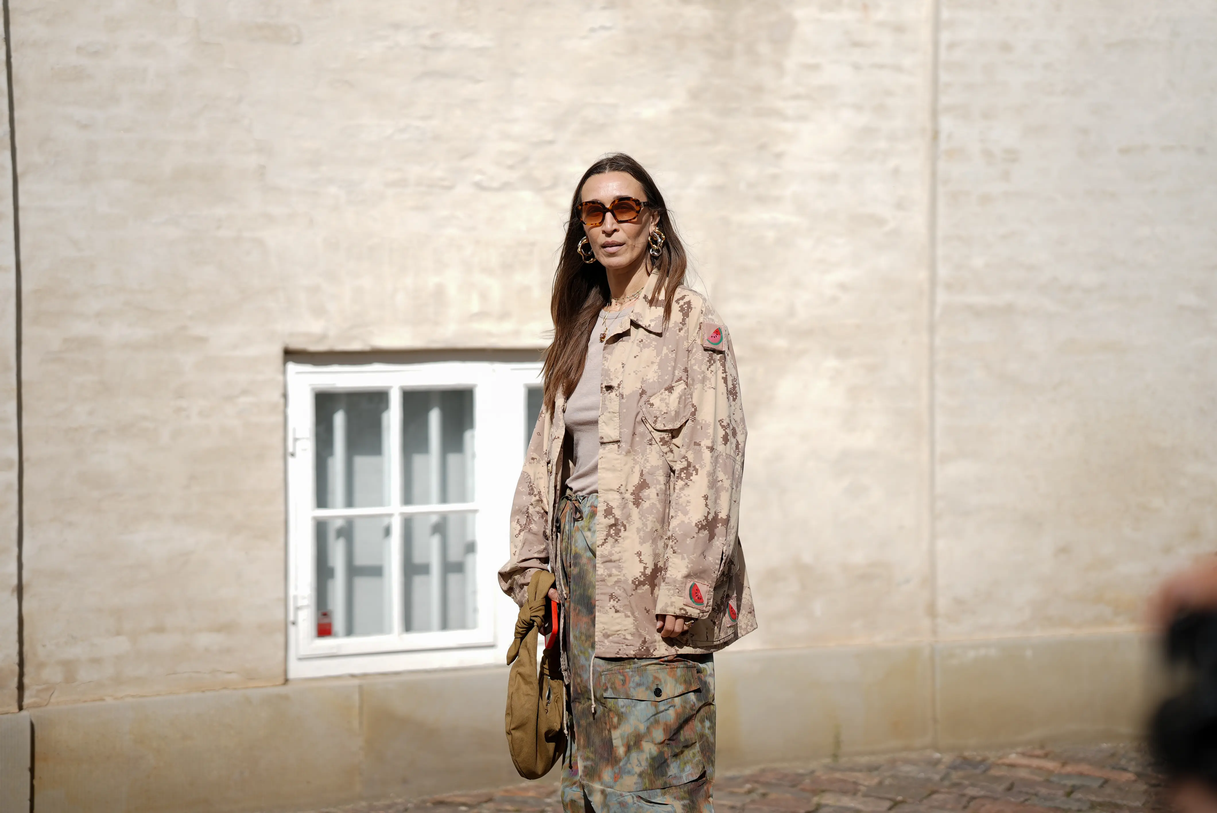COPENHAGEN, DENMARK - AUGUST 5: Alana Hadid, attending the Skall Studio show during Copenhagen Fashion Week SS26, is seen wearing a beige and brown digital camouflage oversized button-up jacket with front flap pockets, long sleeves and small watermelon patches on the sleeves; a taupe crew-neck T-shirt underneath; wide-leg drawstring field cargo trousers in a earth tone colored camouflage design from Henrik Vibskov; a khaki nylon pouch-style handbag with a knotted top handle; square-shaped tortoiseshell sunglasses with rounded glasses; large gold and silver abstract-shaped hoop earrings; her straight brown hair is worn loose with a center part during Copenhagen Fashion Week day two on August 5, 2025 in Copenhagen, Denmark.  (Photo by Moritz Scholz/Getty Images)