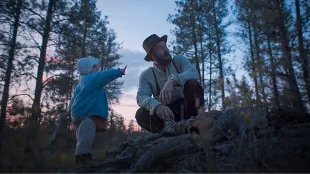 Robert Grainier kneels beside young Katie in the Idaho woods at dusk in Train Dreams Image via Netflix.