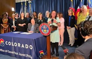 Colorado State Senator Faith Winter standing on the podium beside Governor Jared Polis (Image via Facebook/Senator Faith Winter)